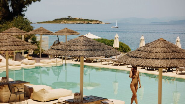Área de piscina com espreguiçadeiras e guarda-sóis de palha, com vista para um corpo d'água onde se vê um veleiro ao longe.