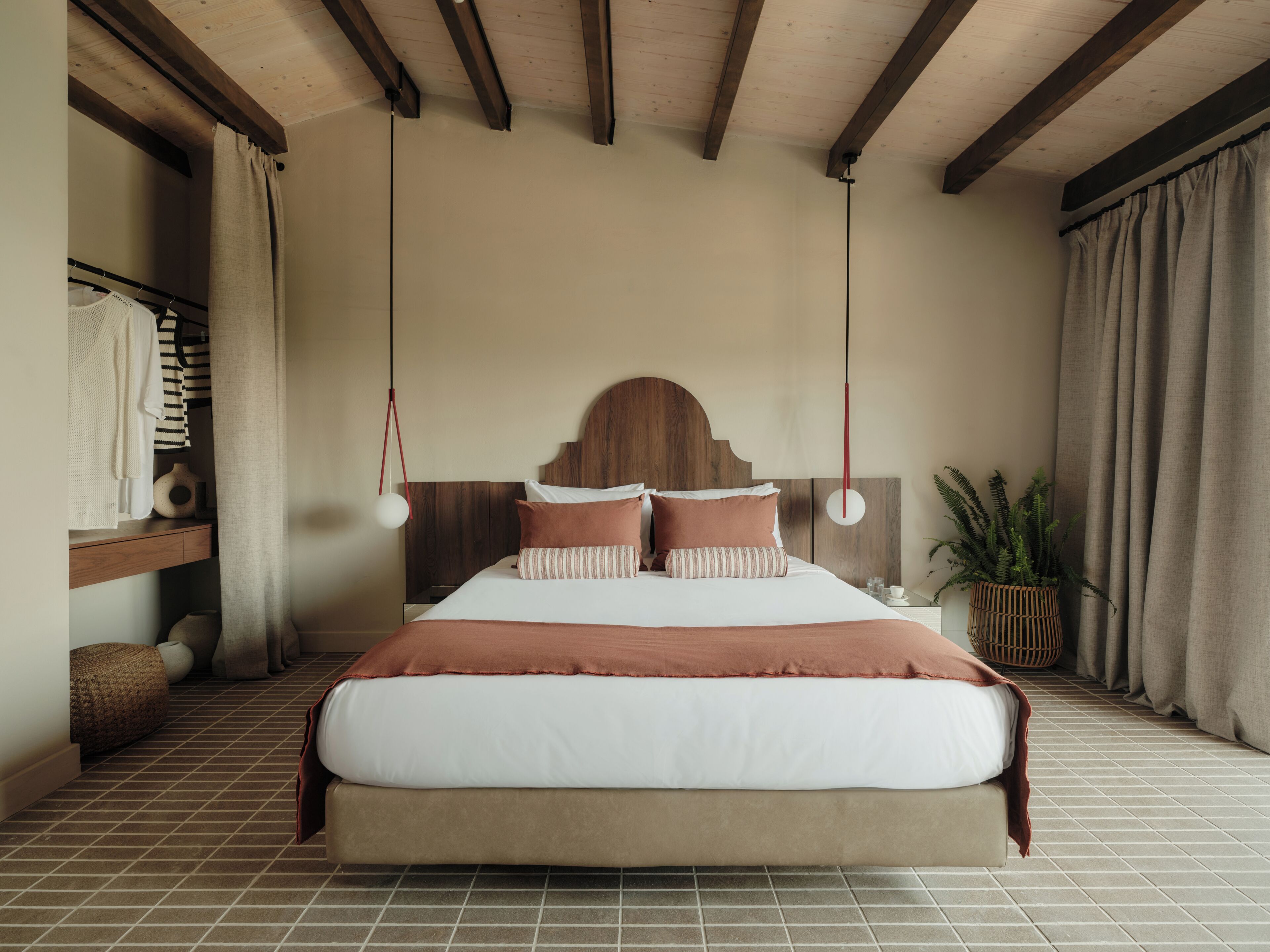 Ein Schlafzimmer mit einem großen Bett, einem Holz-Kopfteil, hängenden Pendelleuchten, einem Nachttisch, einer Topfpflanze und Blick ins Freie.