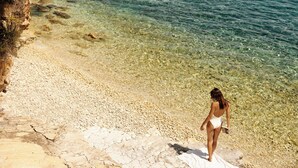 Uma mulher de fato de banho caminhando por uma praia rochosa com água cristalina.