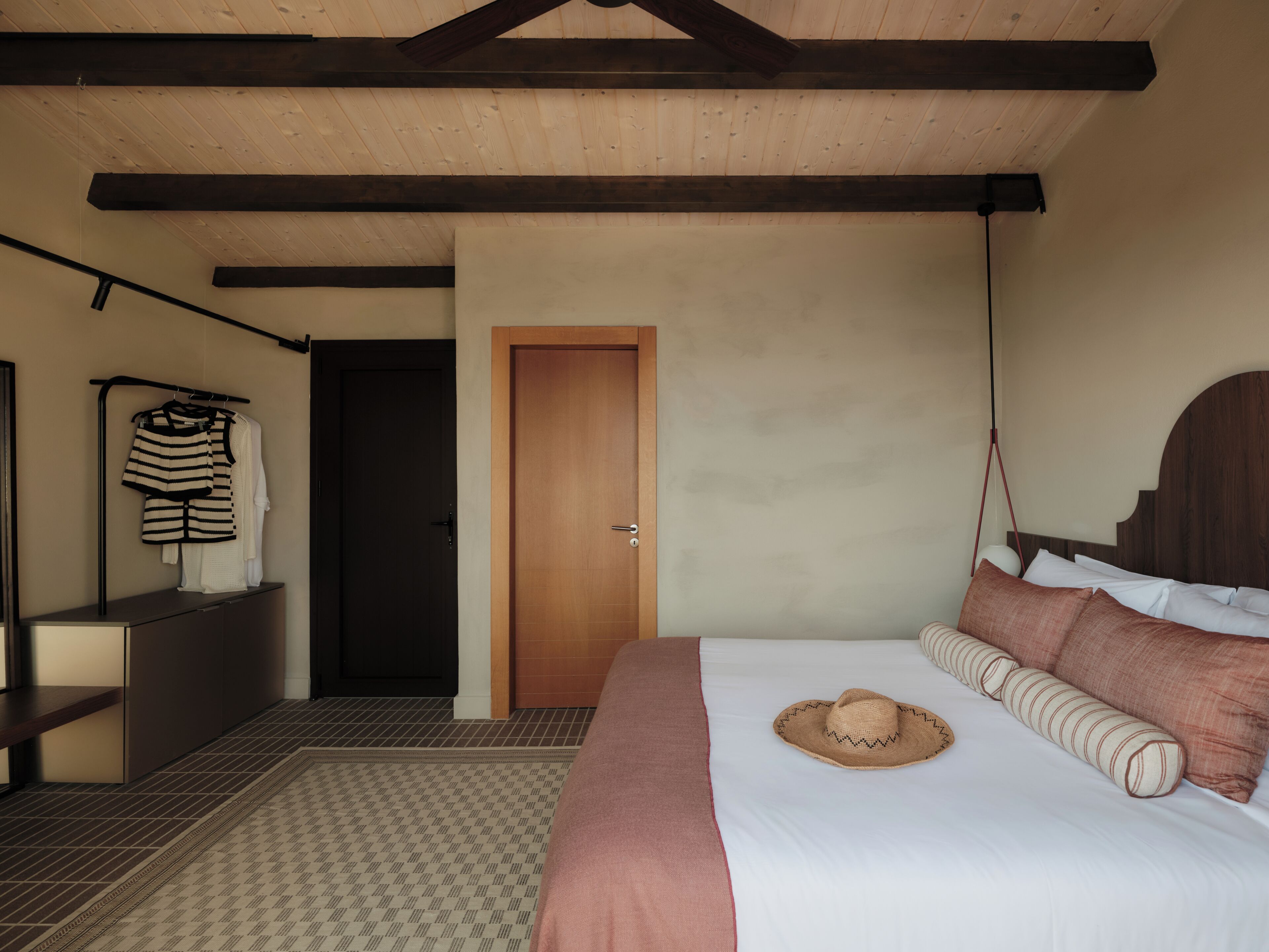A bedroom with a bed, a wooden ceiling, a door, and a clothing rack.