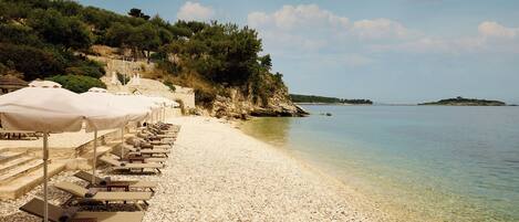 A pebbled beach with sun loungers and umbrellas, a clear view of the sea, and a rocky hillside with trees.