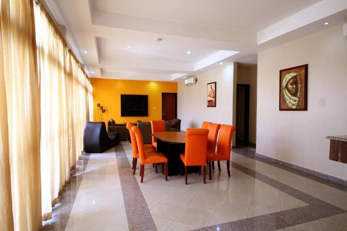 A hotel lobby with a round table and orange chairs, a flat-screen TV, and a large window with curtains.