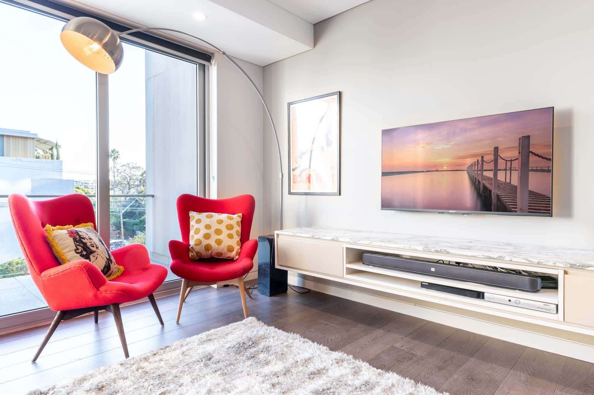A modern living room with a large window, a red armchair, a white sofa, a flat-screen TV, and a framed picture of a sunset.