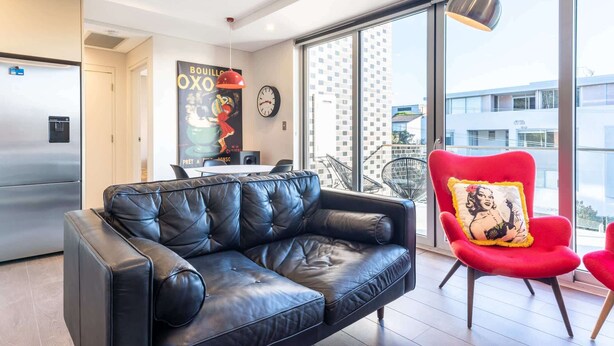 Un salon moderne avec un canapé en cuir noir, un fauteuil rouge agrémenté d’un coussin décoratif, une table à manger avec des chaises, une grande fenêtre donnant sur des immeubles et un réfrigérateur.