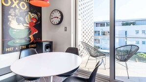 Un espace de salle à manger moderne avec une table blanche, des chaises noires, un luminaire suspendu rouge, une grande affiche, une horloge murale et une vue sur le paysage urbain à travers de grandes fenêtres.