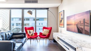 Un salon moderne avec un canapé en cuir, deux fauteuils rouges ornés de coussins à motifs, une cheminée en marbre et de grandes fenêtres offrant une vue sur les bâtiments et le coucher de soleil.