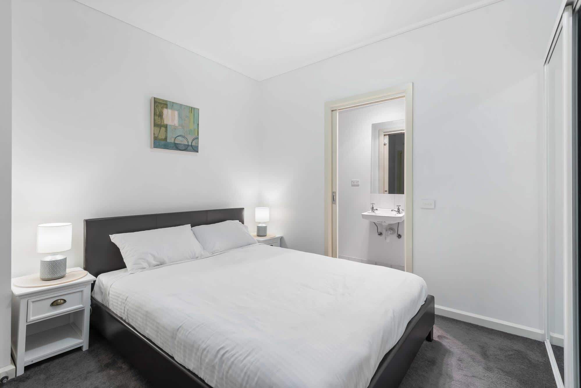 Ein ordentlich eingerichtetes Schlafzimmer mit Bett, Nachttischen, einem Gemälde und einem Badezimmer mit Waschbecken und Spiegel.