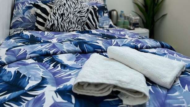 Ein ordentlich bezogenes Bett mit einer blau-weißen blumigen Tagesdecke, einem zebra-gemusterten Kopfkissen und zwei gefalteten weißen Handtüchern. An der Wand hängen zwei abstrakte Gemälde und rechts steht eine Stehlampe.