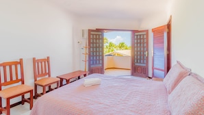 A bedroom with a bed, two wooden chairs, a small table, and a window with a view of palm trees.