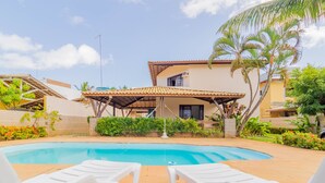 A swimming pool with white lounge chairs, a covered patio, and a house with a tiled roof and palm trees.