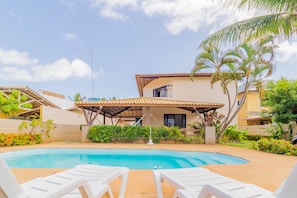Una piscina con sillas de descanso blancas, un porche cubierto y una casa con techo de tejas y palmeras.