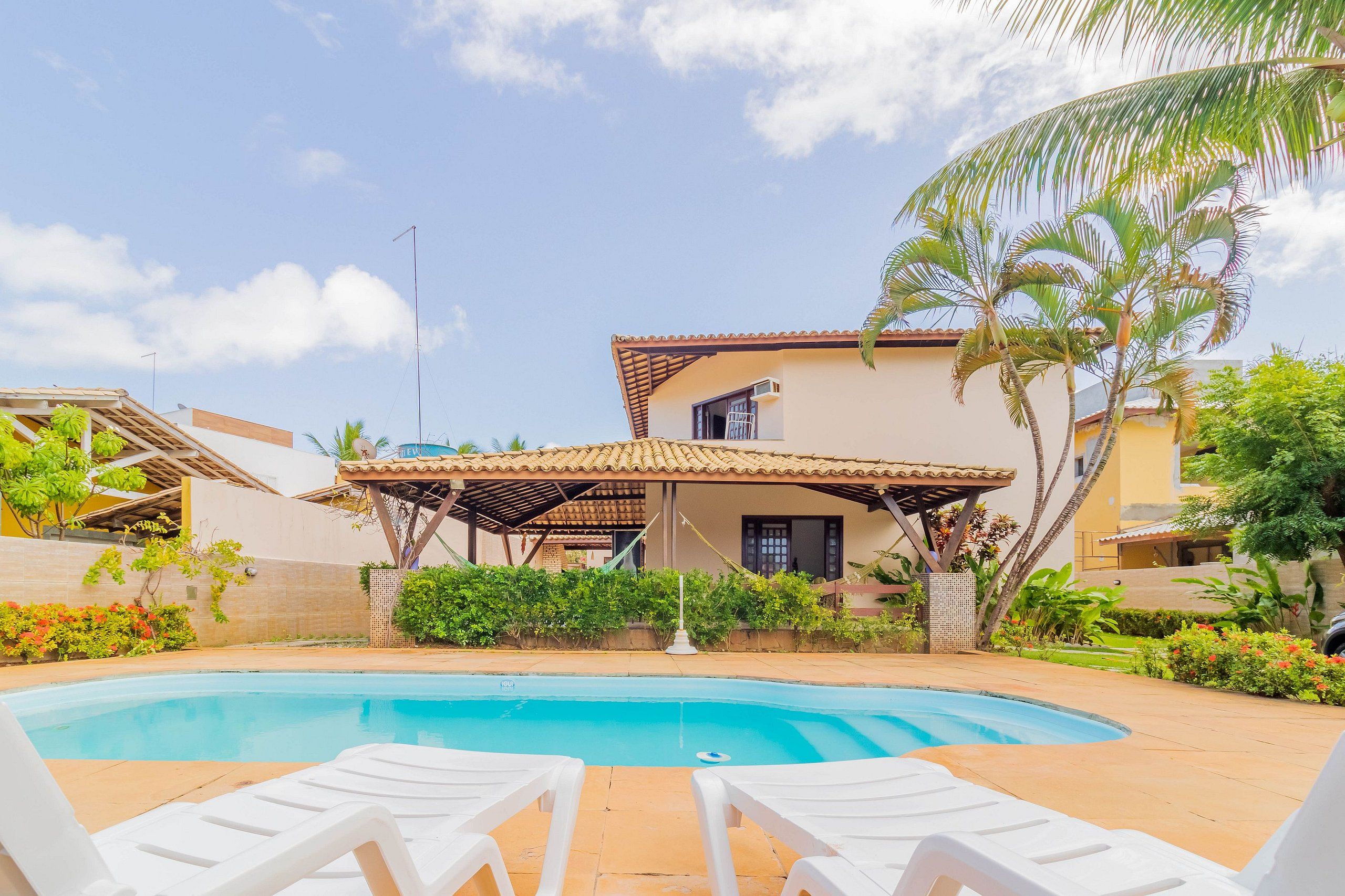 Ein Poolbereich mit zwei weißen Liegestühlen, einer überdachten Terrasse und einem Haus mit einem Ziegeldach.