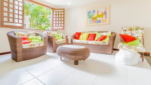 A room with wicker furniture, a white ottoman, and a large window with a view of greenery.