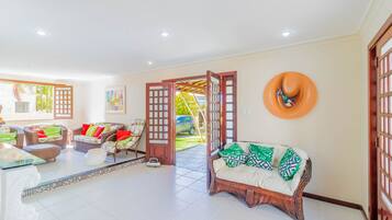 Amplia habitación con un gran ventanal abierto que da al exterior, un sofá de mimbre con cojines, una mesa de centro de cristal y un objeto naranja fijado a la pared.