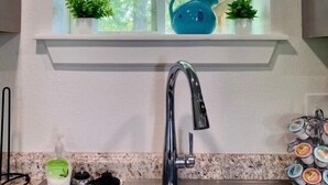 A kitchen with a double stainless steel sink, a window above the sink with two small potted plants, and a teal kettle on the windowsill. - Swim, fish, and explore bluffs, just across the street. (Lowell)