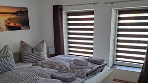 A modern bedroom with a large bed, wooden flooring, and a view of the outdoors through large windows with blinds. - Speicher-studio 3 (Klein Upahl)