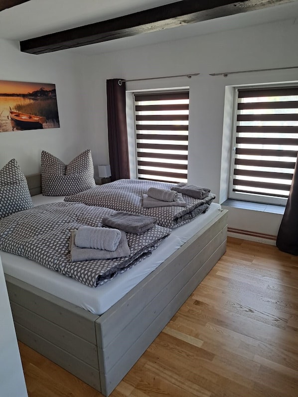 Une chambre moderne avec un grand lit, un plancher en bois et une vue sur l’extérieur grâce à de grandes fenêtres équipées de stores.