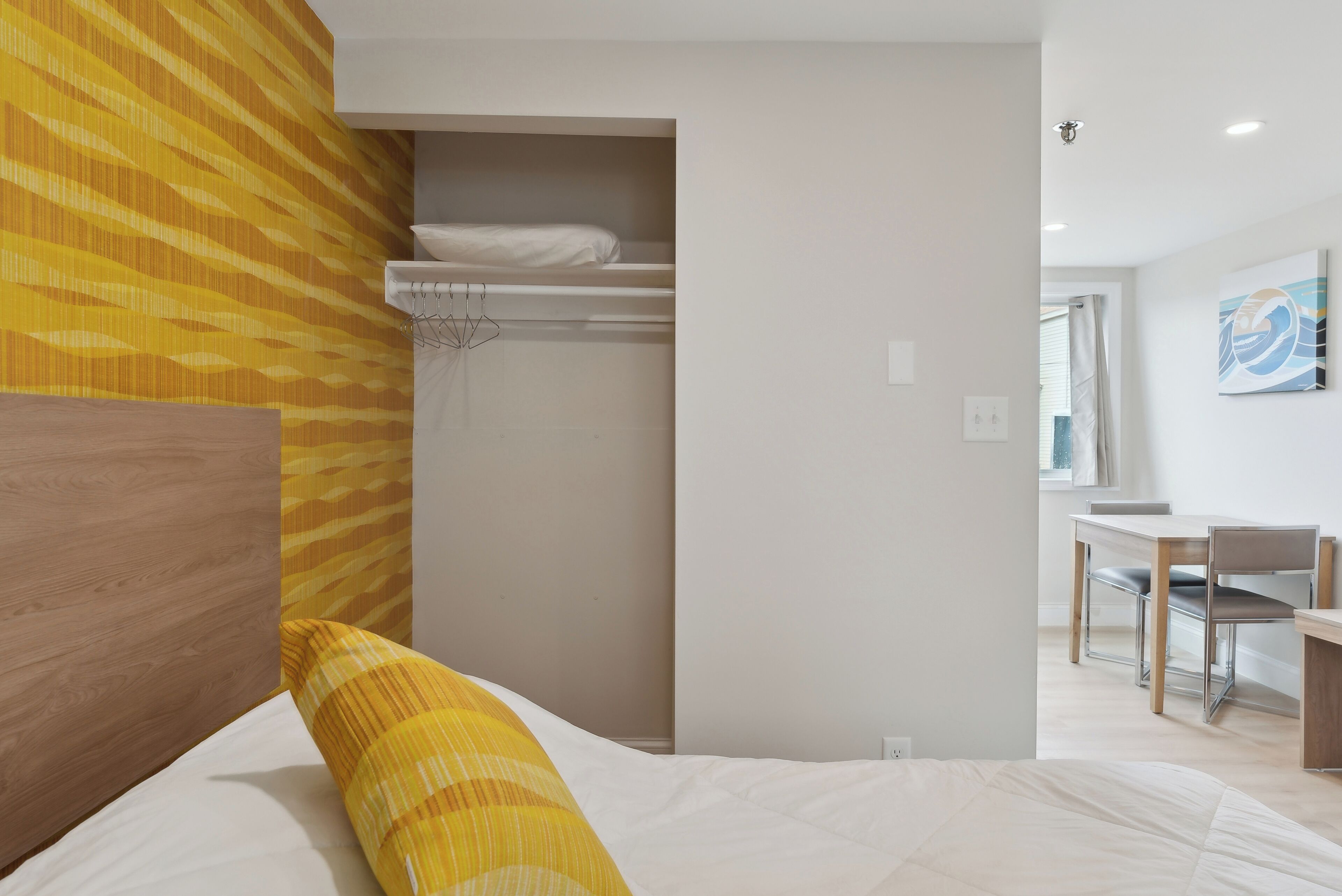 A bedroom with a bed, a wooden headboard, a yellow-striped pillow, a white bedspread, a built-in shelf with a white mattress, and a desk with a chair in the adjacent room.