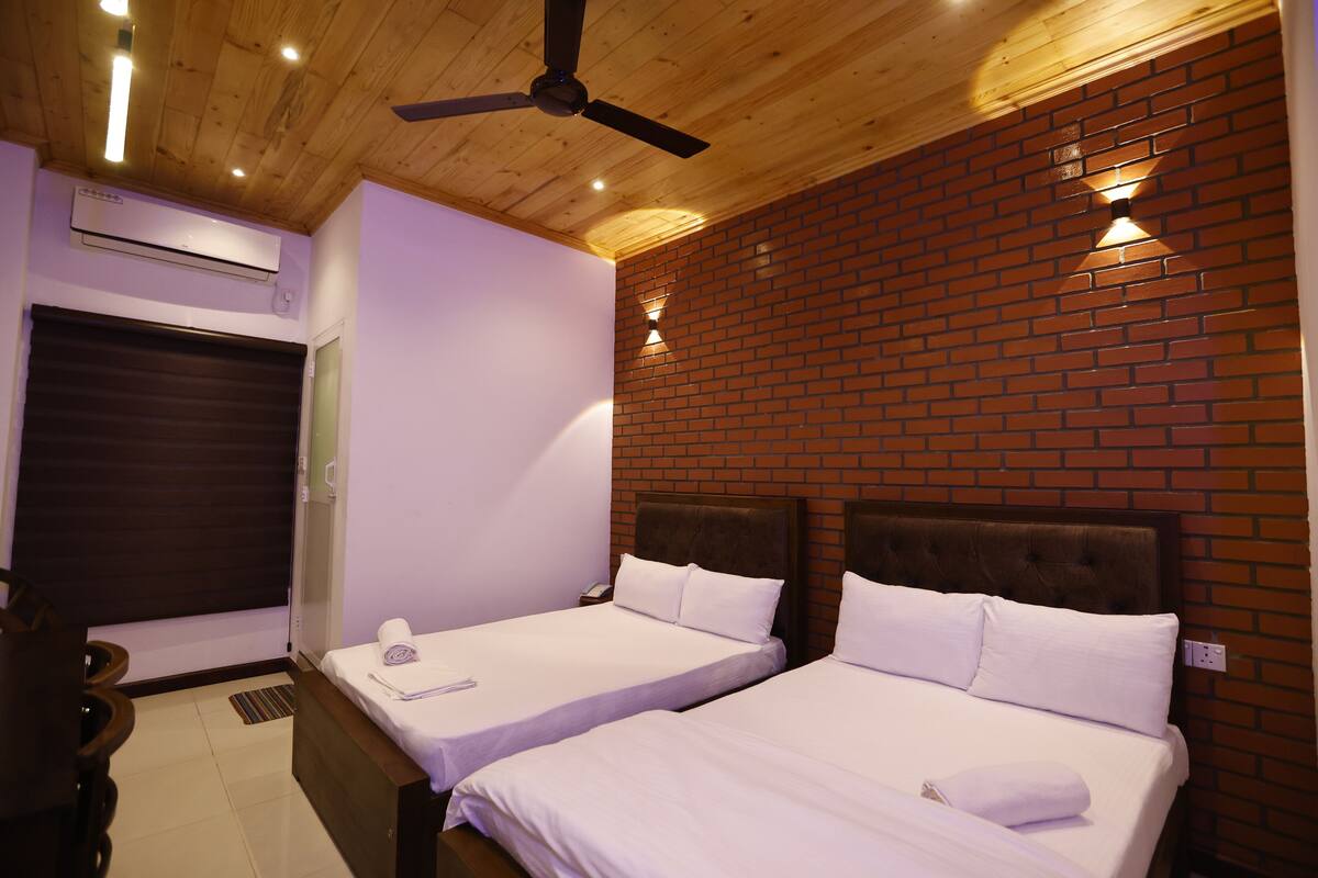 A hotel room with two beds, a ceiling fan, a brick wall, and a window with a closed shutter.