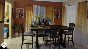 Ein Essbereich mit Tisch, Stühlen und einer Vase mit Blumen. An der Wand hängen zwei Gemälde und ein Fenster mit Vorhängen.