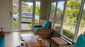 A room with a large sliding glass door leading to a driveway, wicker furniture with teal cushions, and a wooden coffee table. - Island Style Villa in Richmond Estate Palms (St. Ann Parish)