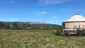 Une yourte se dresse dans un champ verdoyant, sous un ciel dégagé, avec des collines qui se dessinent à l’horizon.