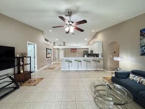 A modern living room with a TV, a glass coffee table, a sofa, a ceiling fan, and a kitchen area in the background.