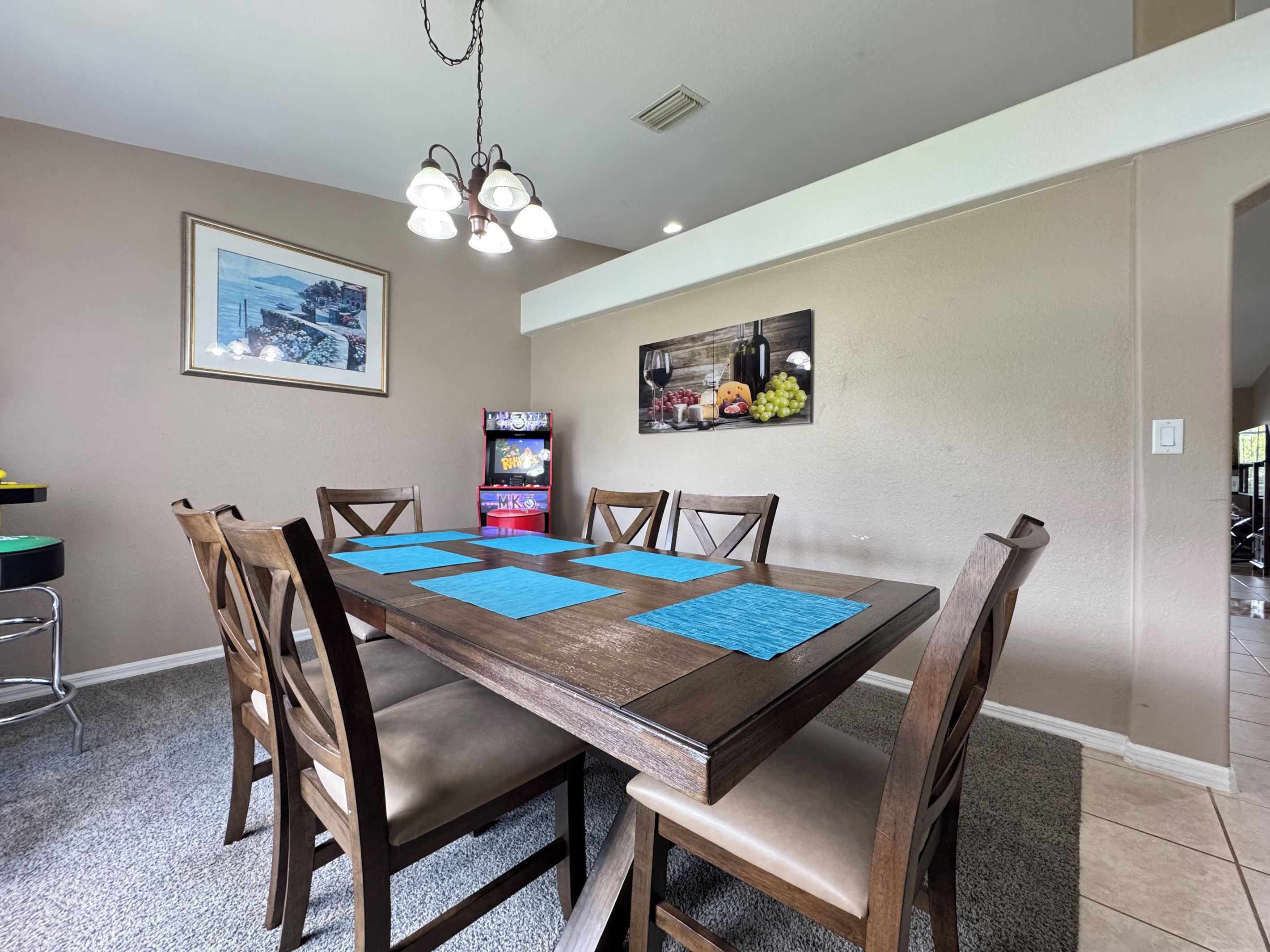 Un coin repas avec une table en bois, des chaises, des sets de table bleus, un lustre, des œuvres d’art mural et une console de jeux vidéo.