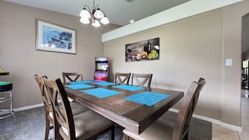 Uma sala de jantar com mesa de madeira, cadeiras, jogos americanos azuis, um candelabro, obras de arte nas paredes e um console de videogame.