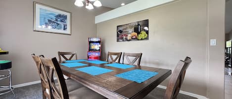 Un coin repas avec une table en bois, des chaises, des sets de table bleus, un lustre, des œuvres d’art mural et une console de jeux vidéo.