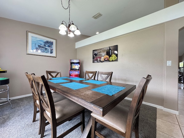 A dining area with a wooden table, chairs, blue placemats, a chandelier, wall art, and a video game console.