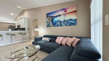 A modern living room with a sofa, a glass coffee table, and a kitchen area in the background.