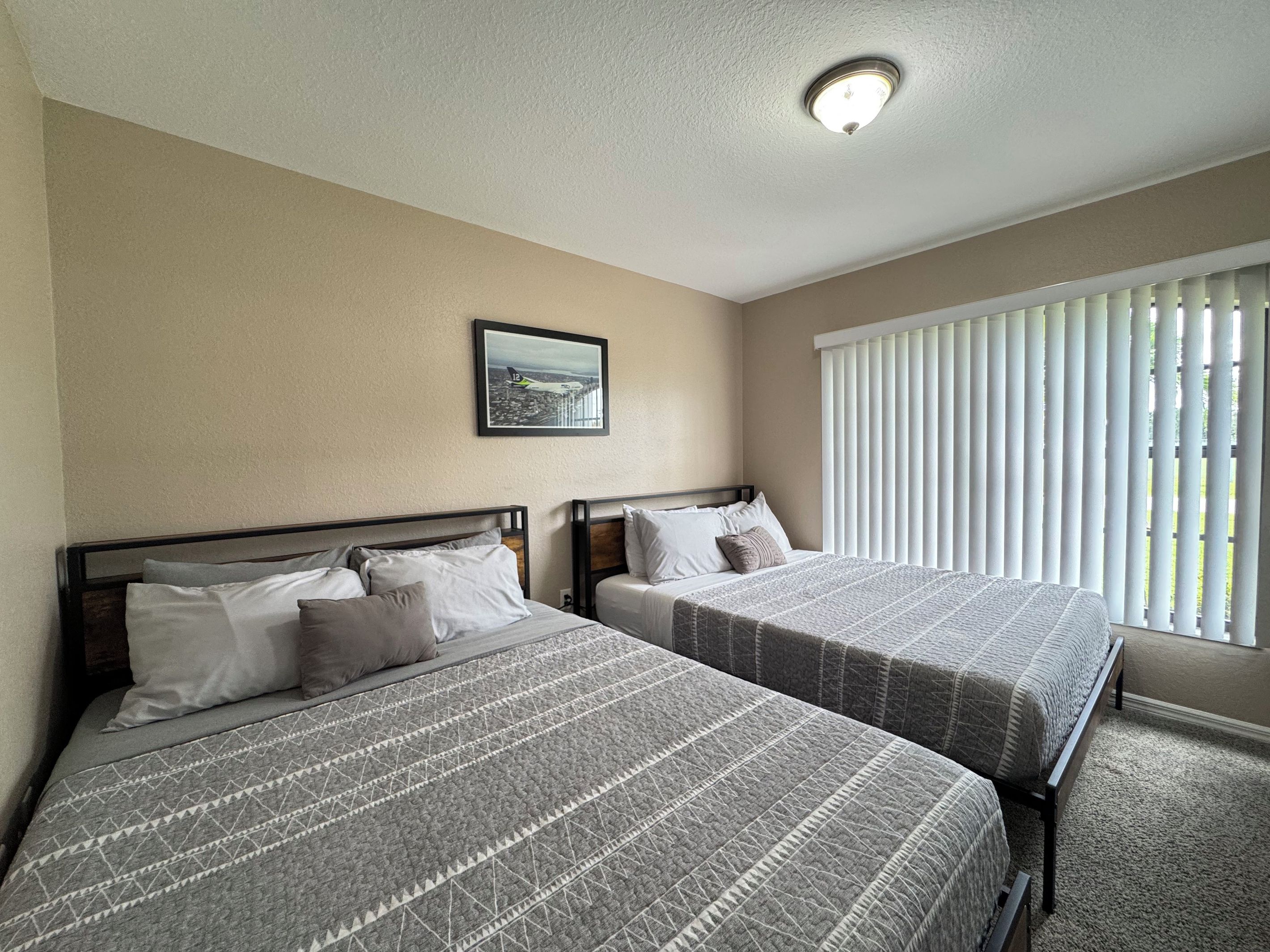 A hotel room with two beds, a ceiling light, a window with blinds, and a framed picture on the wall.