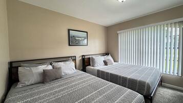 A hotel room with two beds, a ceiling light, a window with blinds, and a framed picture on the wall.
