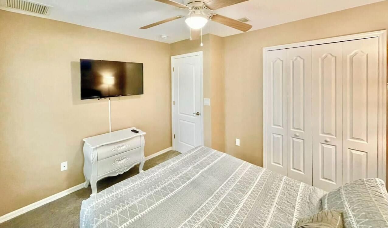Une chambre à coucher comprenant un lit, une commode, un téléviseur fixé au mur, un placard et un ventilateur de plafond.