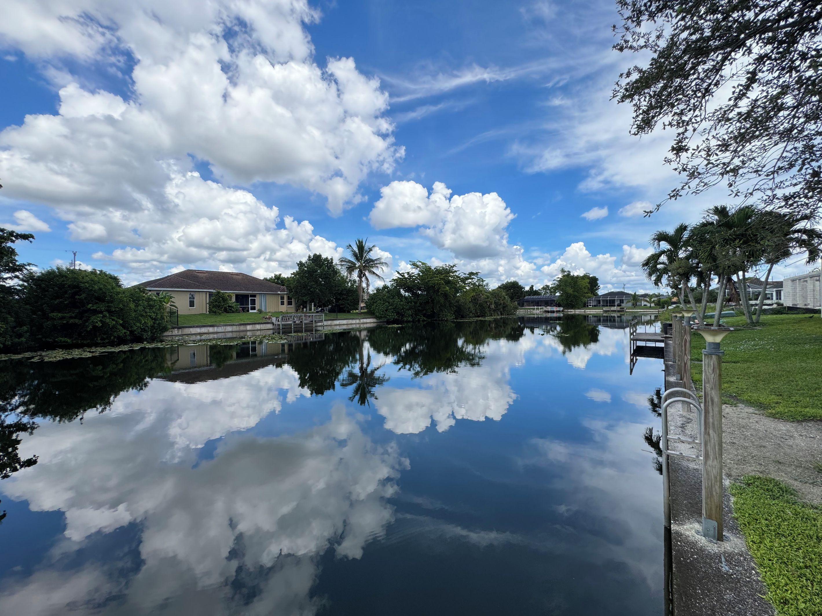 A calm water body reflecting the sky and surrounding buildings, with a clear view of a house and palm trees.