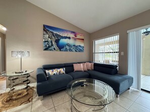 A living room with a dark sofa, a glass coffee table, a painting of a mountainous landscape, and a lamp.