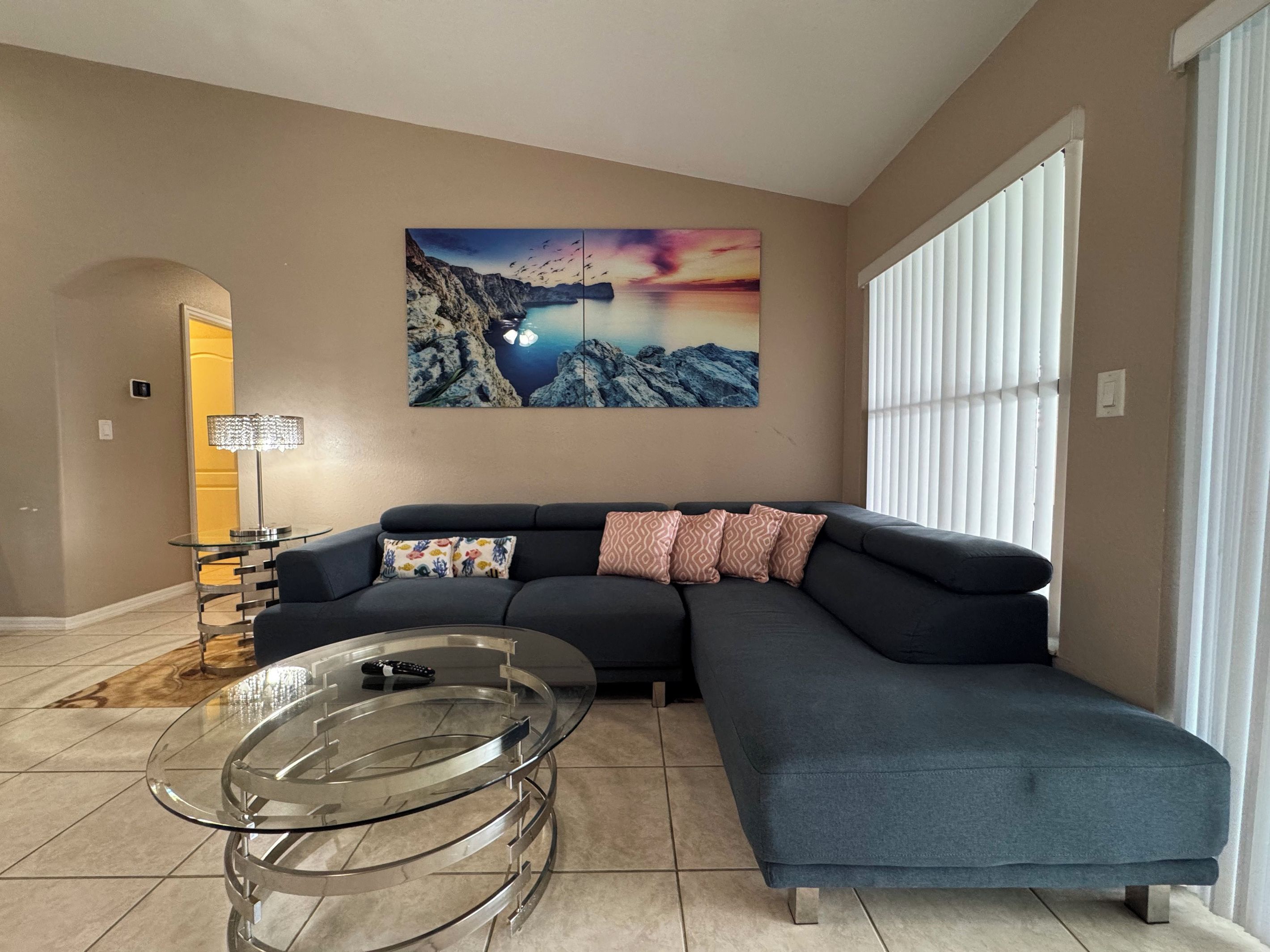 A living room with a sectional sofa, a glass coffee table, and a large landscape painting on the wall.