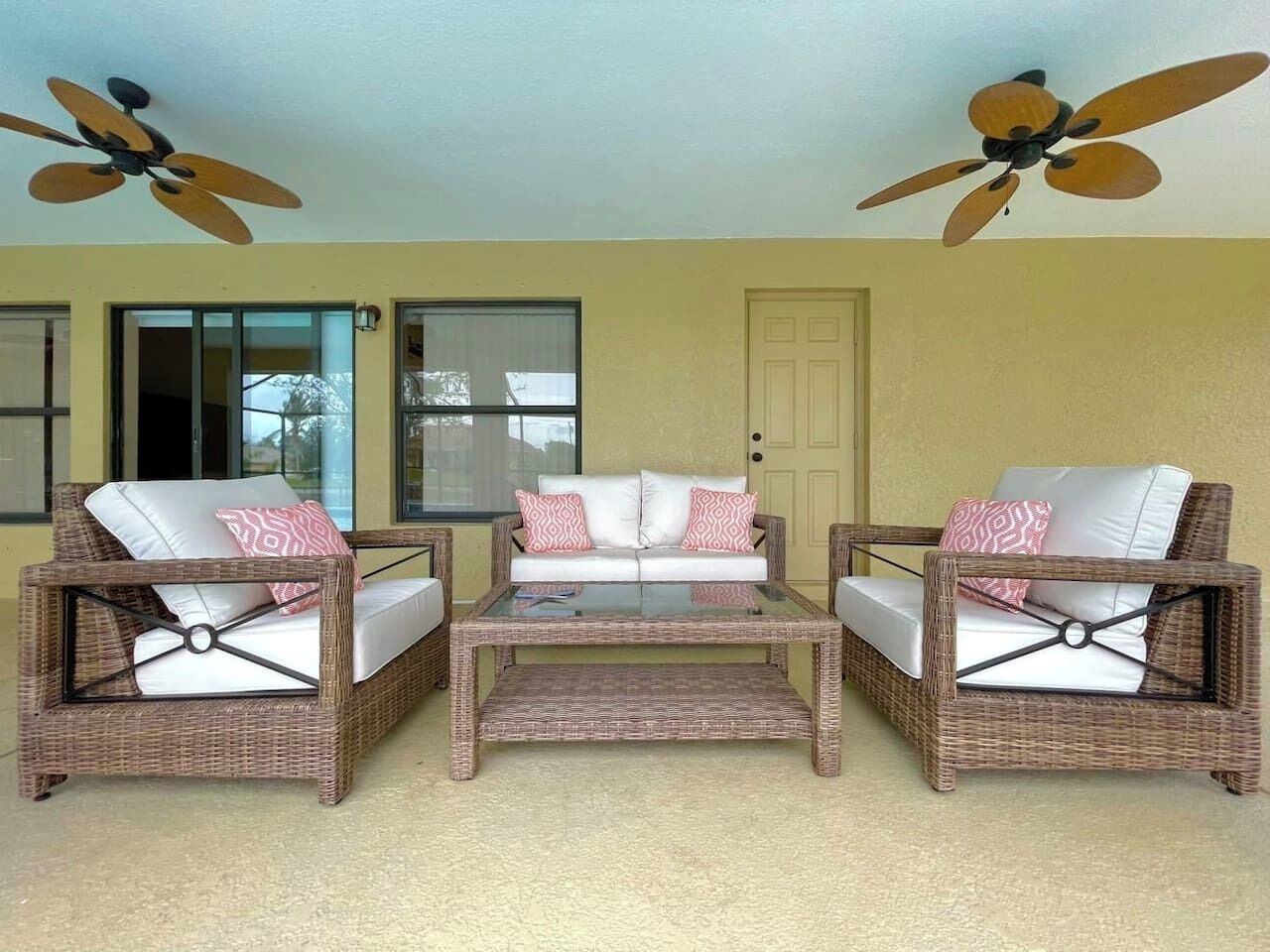 A room with wicker furniture, including a sofa and two armchairs, a glass coffee table, and ceiling fans.
