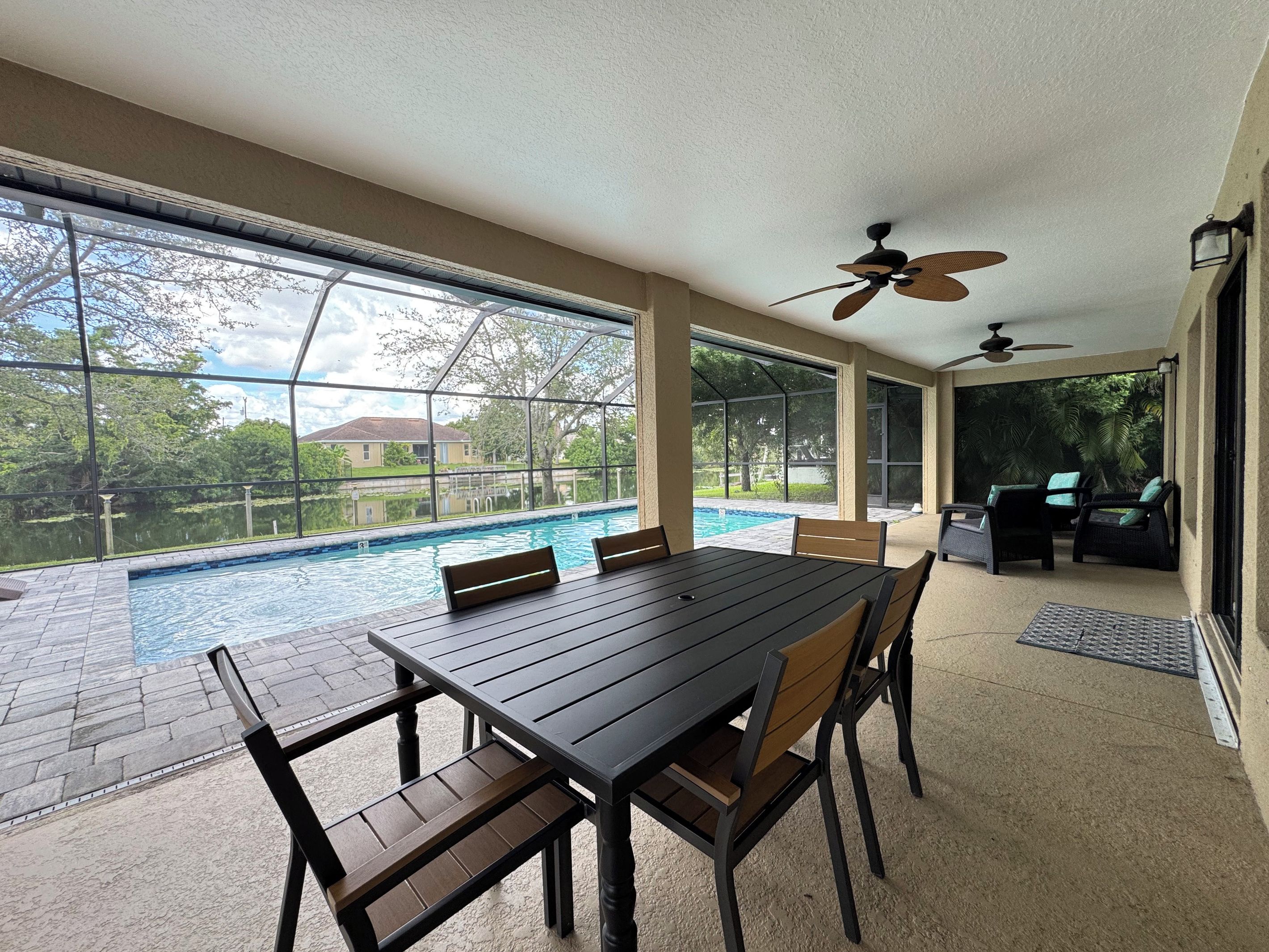A poolside dining area with a table and chairs, a ceiling fan, and a view of a swimming pool and greenery.