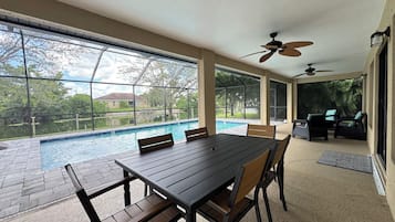 A poolside dining area with a table and chairs, a ceiling fan, and a view of a swimming pool and greenery.