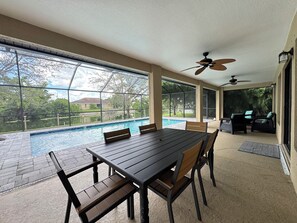 Un área de comedor junto a la piscina con mesa y sillas, ventilador de techo y vista a una piscina y follaje.