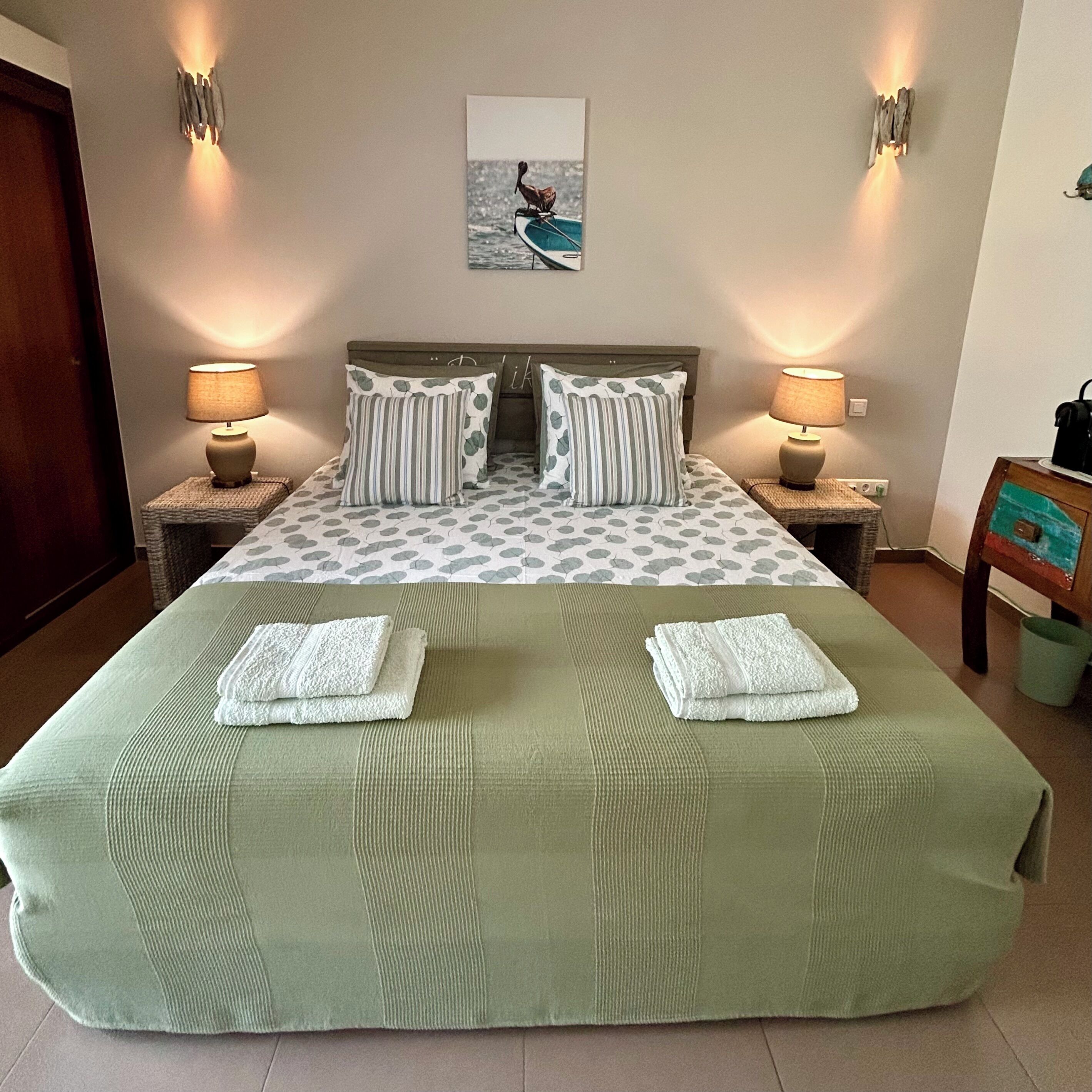 Een tweepersoonsbed met een sprei met bladprint, twee kussens, twee nachtlampjes en een lijst met een afbeelding aan de muur.