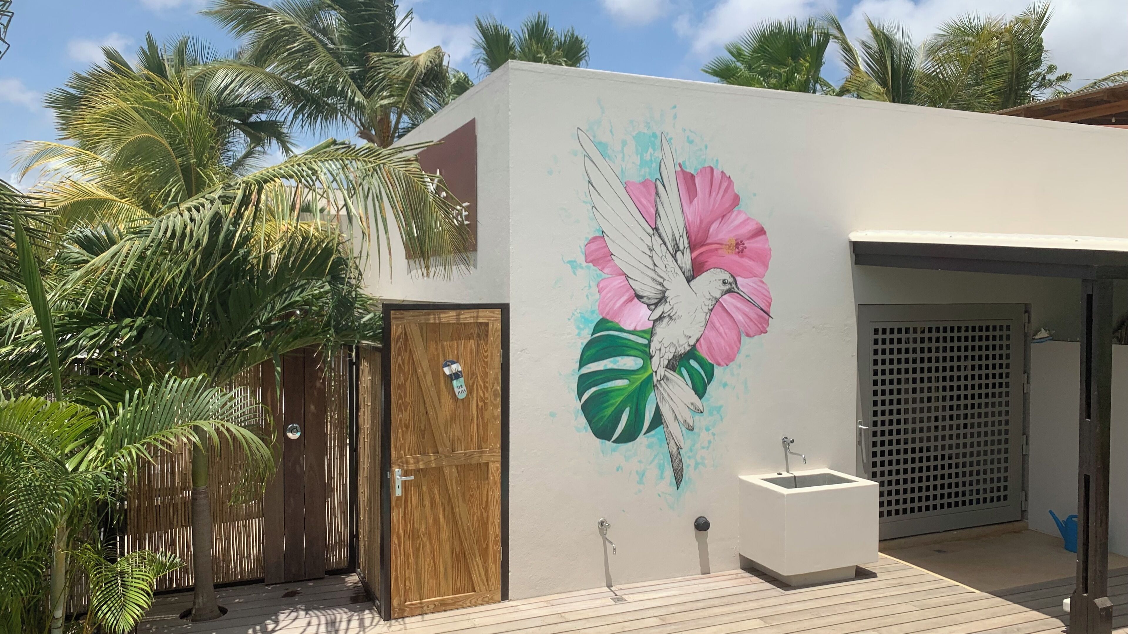 Een modern gebouw met een houten deur, een witte muur met een muurschildering van een vogel en bloemen, een klein buitenwasbakje en een houten terras.