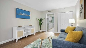 A modern living room with a flat-screen TV, a sofa with yellow cushions, a white entertainment unit, and a potted plant.