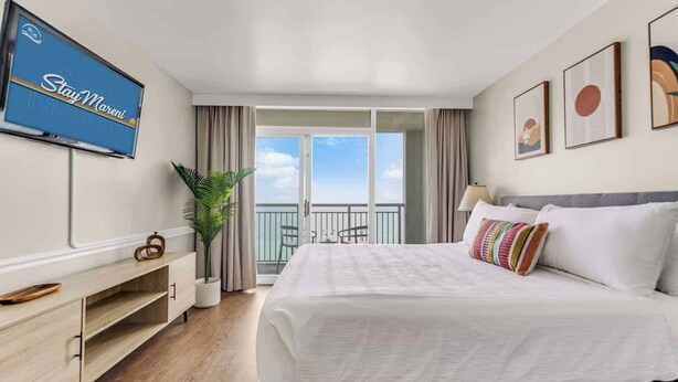 A hotel room with a large bed, a TV on the wall, a balcony with a view, and a plant.