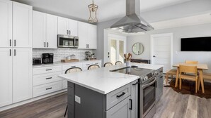A modern kitchen with white cabinets, a central island, stainless steel appliances, and a dining area with a wooden table and chairs. - Palmetto Grove,  Walk to Shem Creek, Mins to Dwt (Mount Pleasant)
