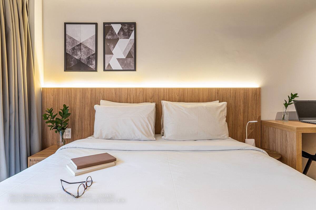Un lit bien fait, avec du linge de lit blanc, une table de chevet sur laquelle se trouve un ordinateur portable, et deux tableaux abstraits accrochés au mur.