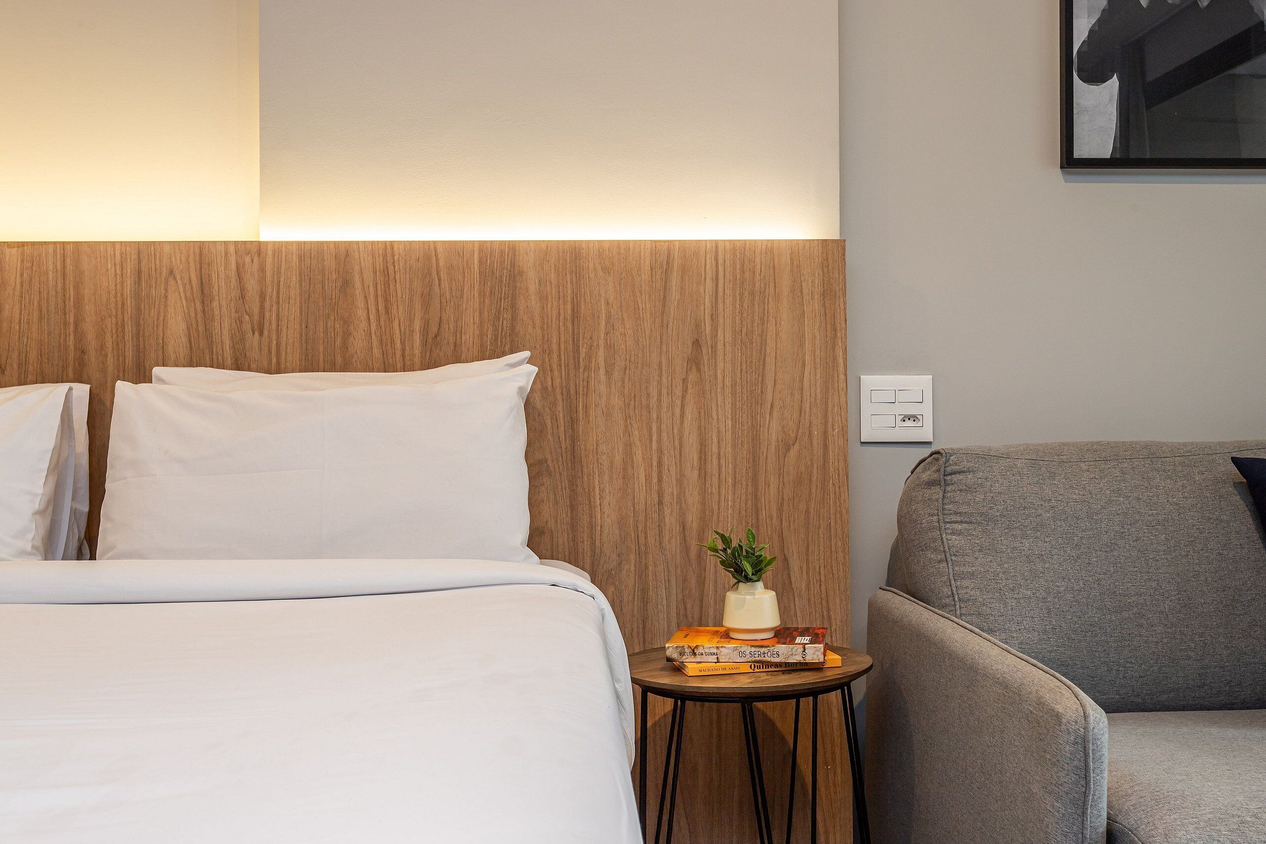 Habitación de hotel con una cama, un sillón gris, una mesita con una planta y libros, y una lámpara de pared.