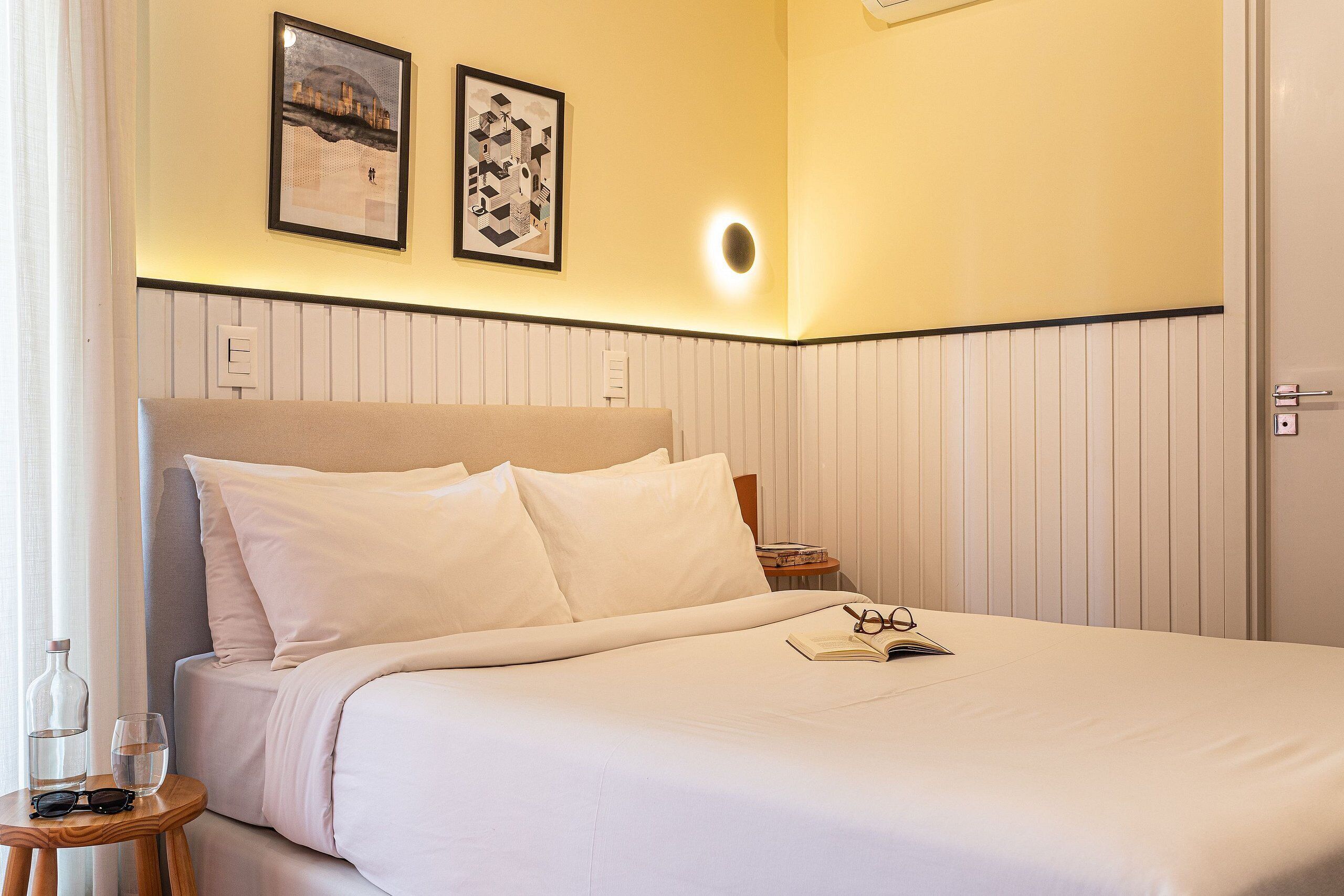 A neatly made bed with pillows, a bedside table with a book and glasses, and two framed pictures on the wall.